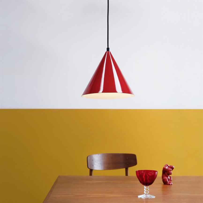 Rød konisk loftslampe hænger og lyser over et træbord; foran en gul-hvid todelt væg står en enkel træstol, et rødt vinglas og en lille rød skulptur.