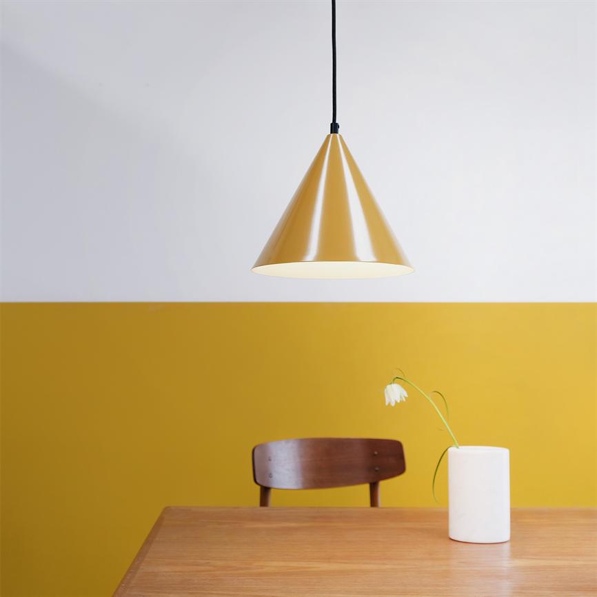 Gul konisk loftslampe hænger over træbord; en træstol står bag bordet, og en enkelt hvid blomst bøjer i en hvid cylinderformet vase; væg er delt i hvid og okkergul.