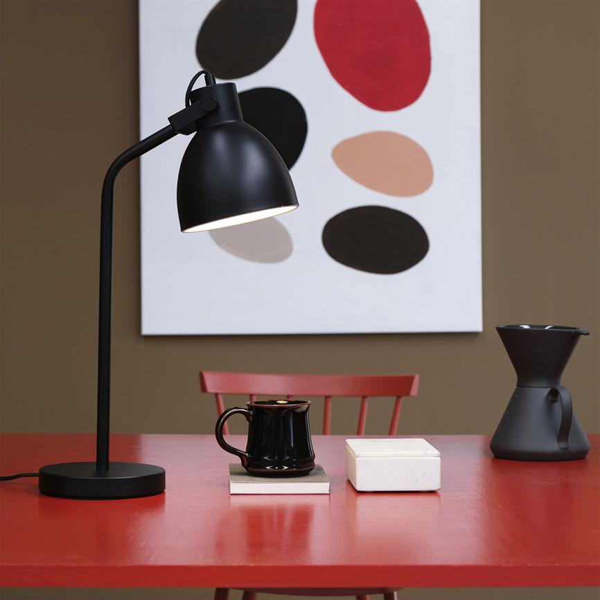 Sort bordlampe lyser ned over en sort krus på en rød bordplade; en rød stol, abstrakt maleri på væggen og en sort kaffebrygger står i baggrunden.
