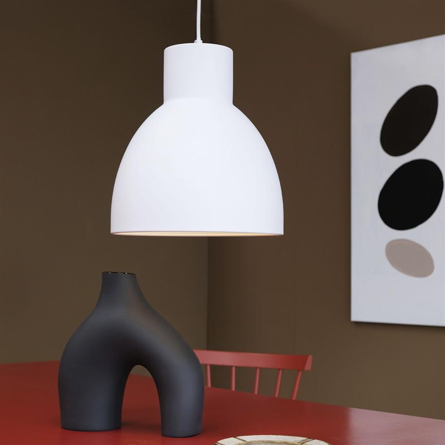 Hvid pendel hænger over et rødt bord med en sort, skulpturel vase, rød stol i baggrunden og en abstrakt plakat på en brun væg.