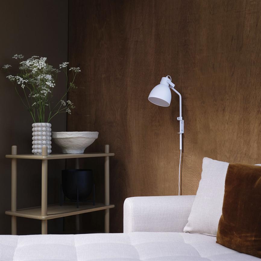 Hvid væglampe er monteret på en varm trævæg over en lysegrå sofa; et lille bord ved siden holder en perlet vase, hvid skål og sort potte med hvide blomster.