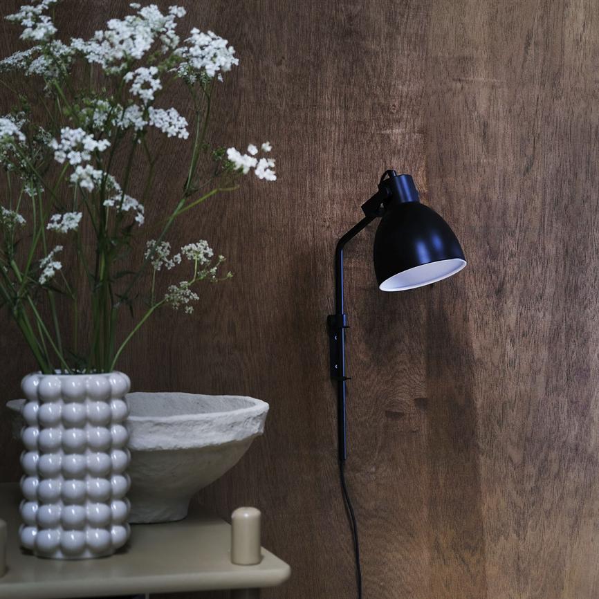 Sort væglampe peger nedad på en mørk trævæg, ved siden af et bord med en hvid perle-tekstureret vase med små hvide blomster og en rå, hvid skål.