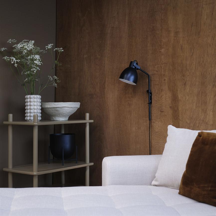 Sort væglampe hænger og peger nedad mod en træbeklædt væg over en lys sofa; ved siden står et lille beige sidebord med hvid vase, skål og sort potte.