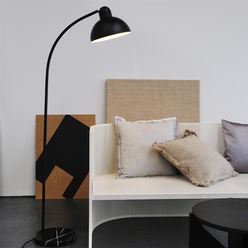 Sort bueformet gulvlampe kaster blødt lys over en hvid net-sofa med tre dekorative puder; bagved står to lærreder lænet mod en lys væg i et minimalistisk, roligt rum.