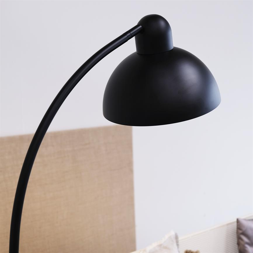 Sort, buet gulvlampe med kuppelformet skærm bøjer sig nedad over en sofa i et lyst rum med en naturfarvet tekstilbagvæg og puder.