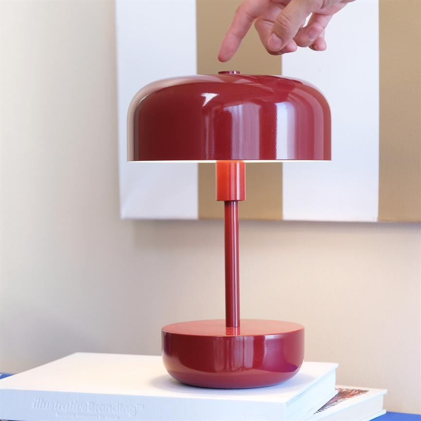 Rød bordlampe med rund skærm; en hånd rækker ned og trykker på knappen øverst; lampen står på en stak hvide bøger foran en lys væg med et maleri. 
 Tekst på bog: "IllustrativeBranding™"