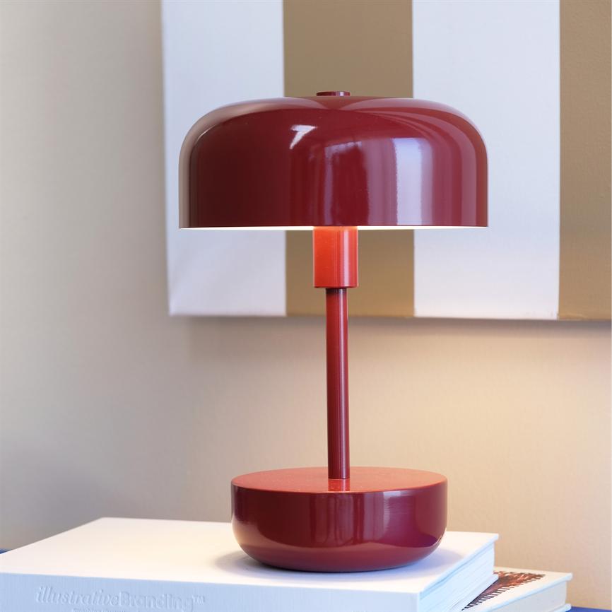 Rød bordlampe lyser blødt; står på en stak bøger foran en beige væg med lodrette hvide og brune striber. Tekst: "illustrativeBranding™".