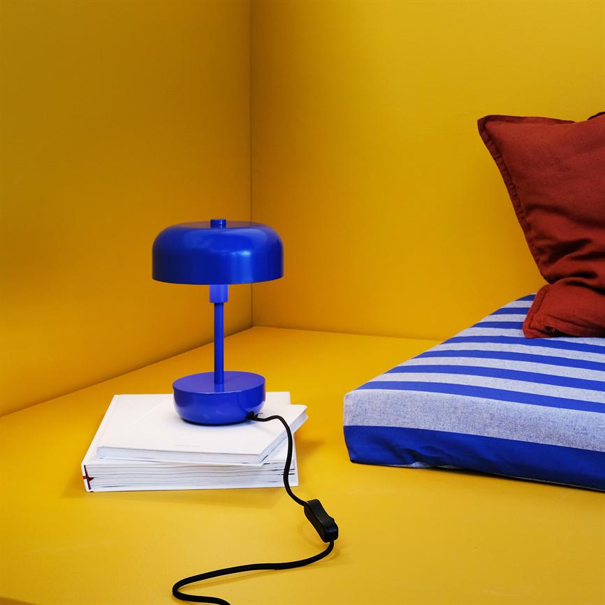Blå bordlampe står på en stak hvide bøger, ledning snor sig over gul flade; ved siden ligger en blå-hvid stribet madras og en rød pude i et gult hjørne.