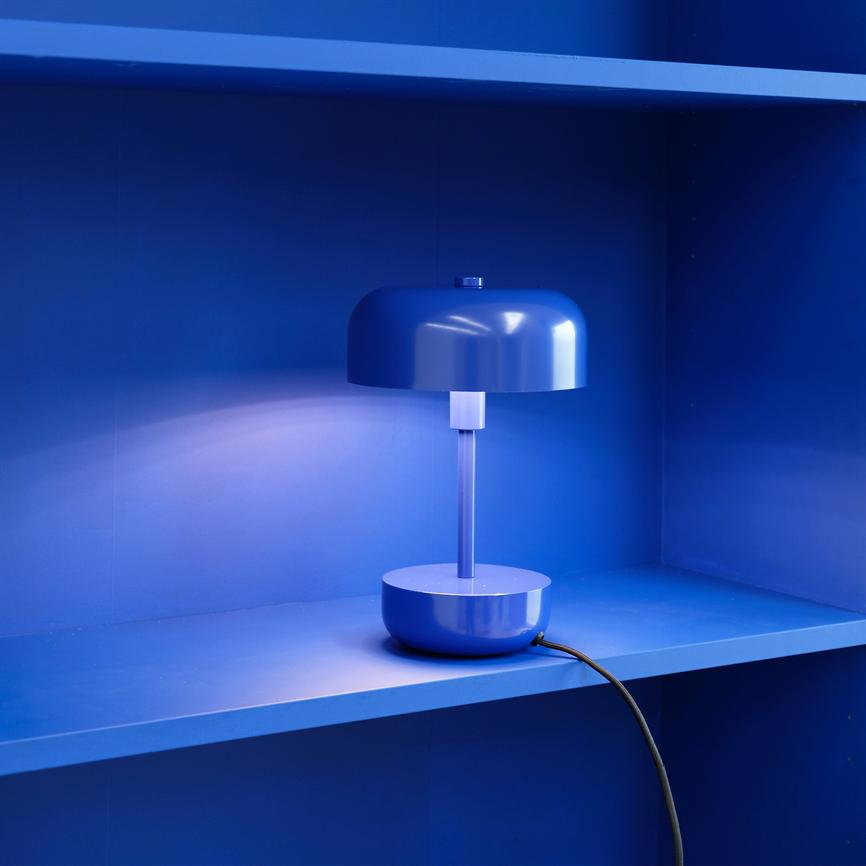 Bordlampe står tændt på en blå hylde i en indbygget blå niche, kaster blødt lys på væggen; en sort ledning løber over hyldekanten.