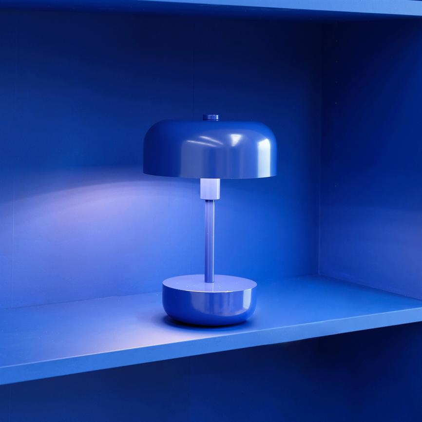 Bordlampe står tændt på en blå hylde i en indrammet niche; den runde skærm kaster blødt lys og omgiver miljøet i intens kobaltblå.