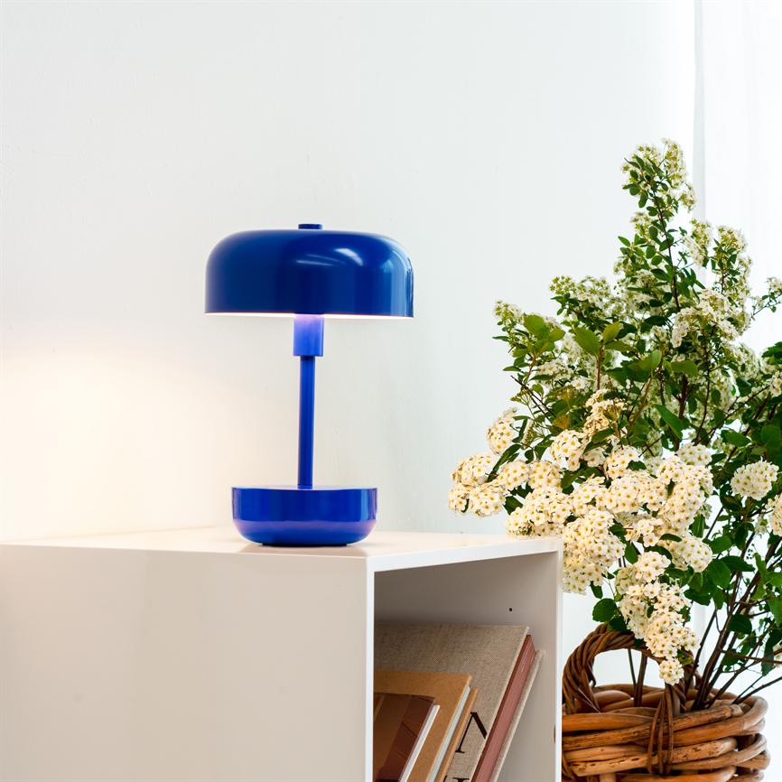 Blå bordlampe lyser svagt over en hvid hylde; ved siden af står en flettet kurv med hvide blomster, mod en lys, neutral væg.