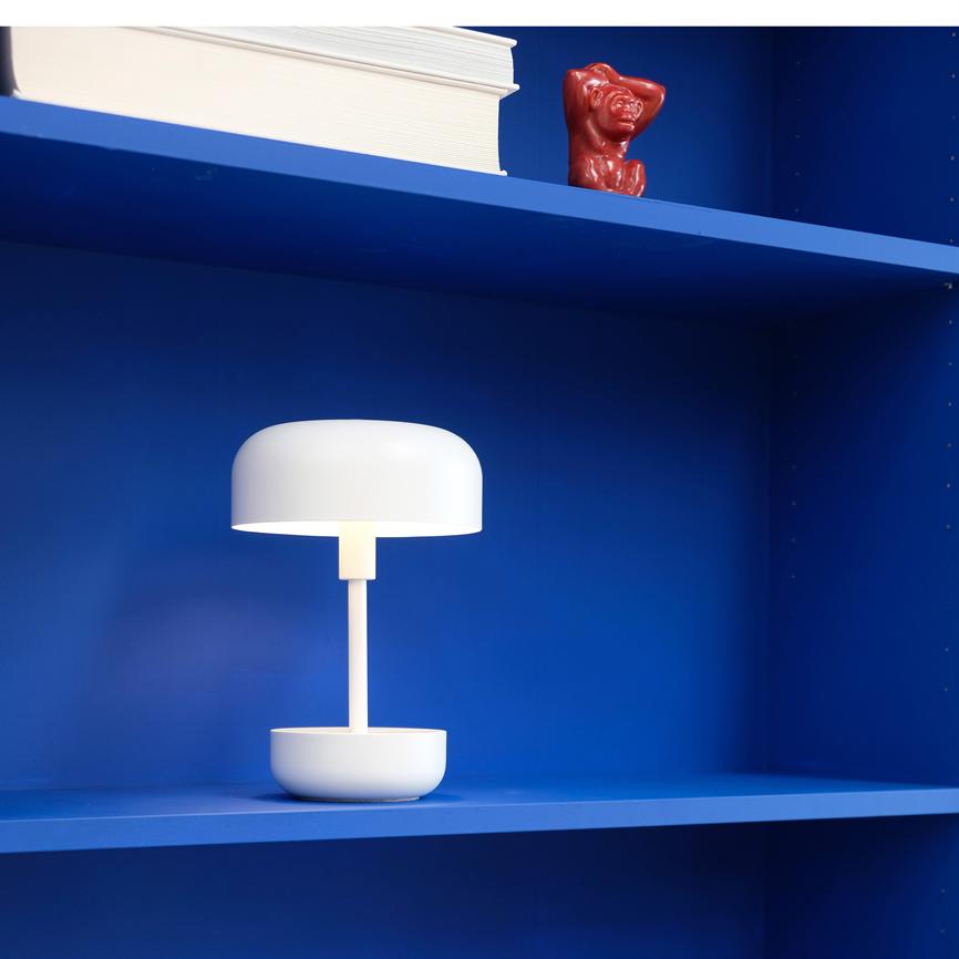 Hvid, moderne bordlampe lyser svagt; står på en klarblå reolhylde med en stak hvide bøger og en lille rød keramikfigur på hylden over.