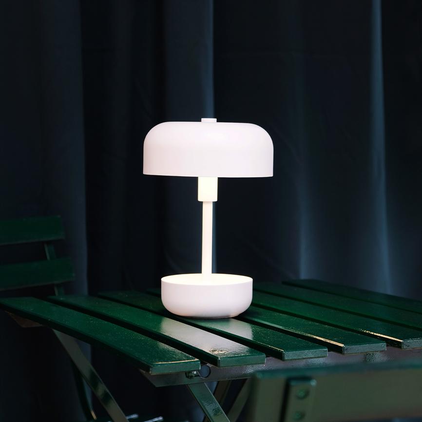 Hvid bordlampe står og udsender blødt, varmt lys på et mørkegrønt sammenklappeligt træbord; baggrunden er mørk med tætte gardiner.
