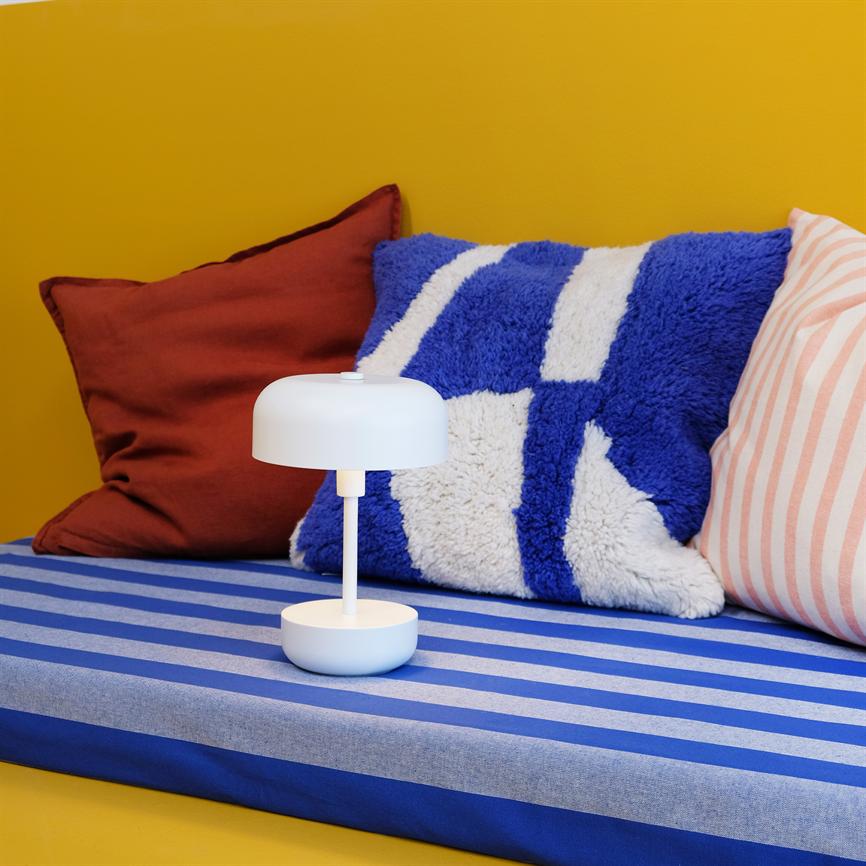 En lille hvid bordlampe står på en blå-lysegrå stribet madras foran en gul væg; omkring den ligger en blå-hvid plyspute, en rustrød pude og en lyserød-hvid stribet pude.