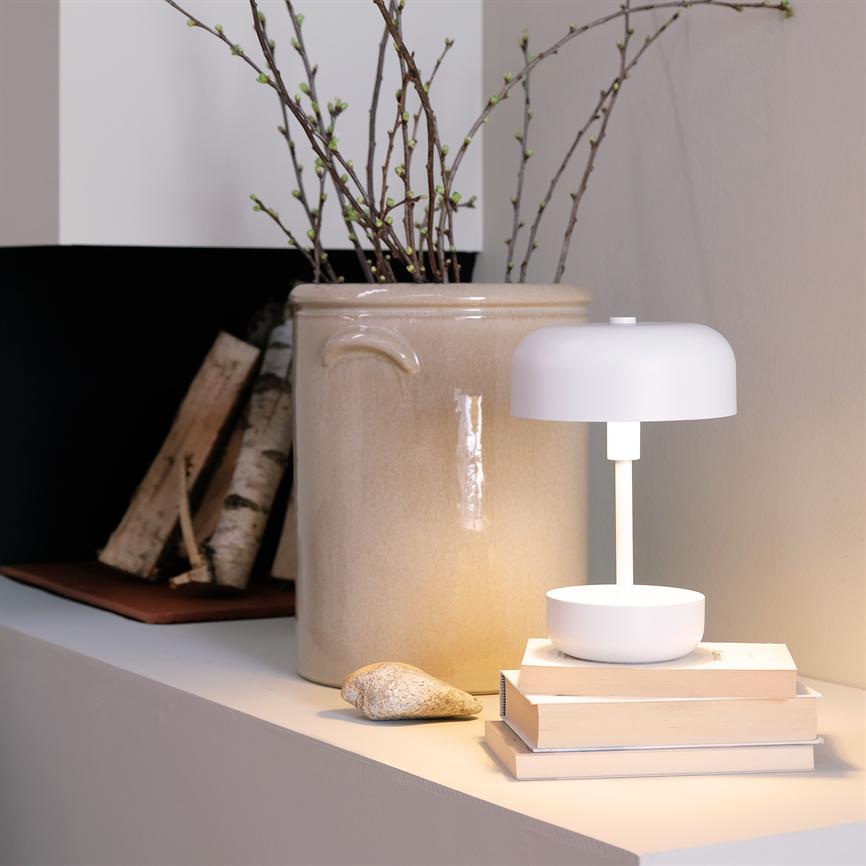 En hvid bordlampe lyser blødt ned på en lille stak bøger; den står på en hylde ved siden af en stor beige krukke med spinkle forårsgrene og birkestammer.