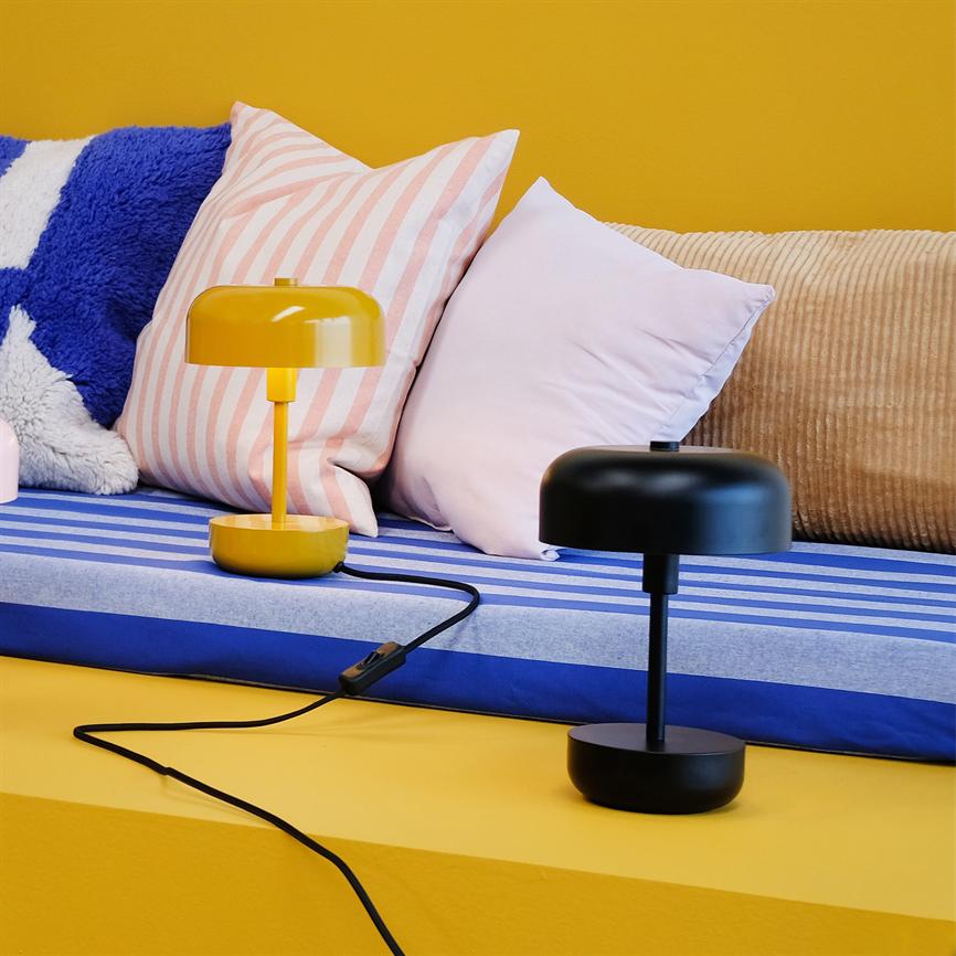 To små lamper, en gul og en sort, står på en blåstribet polstret siddeplads; bløde puder i lyserød, rosa-stribet, blå-hvid og beige lener sig mod en gul væg.
