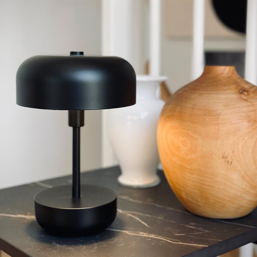 Sort bordlampe står på et mørkt marmorbord, flankeret af en hvid vase og en rund trævase i et lyst, moderne stuehjørne.