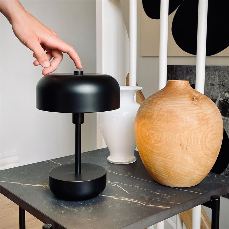 Sort bordlampe, en hånd rækker ud og trykker på dens top; står på et mørkt marmorbord mellem en hvid vase og en rund trævase foran hvide spiler og vægkunst.