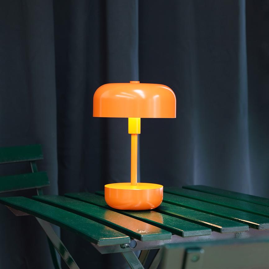 En orange bordlampe med rund skærm lyser blødt og står på et grønt lakeret træbord foran mørke gardiner.