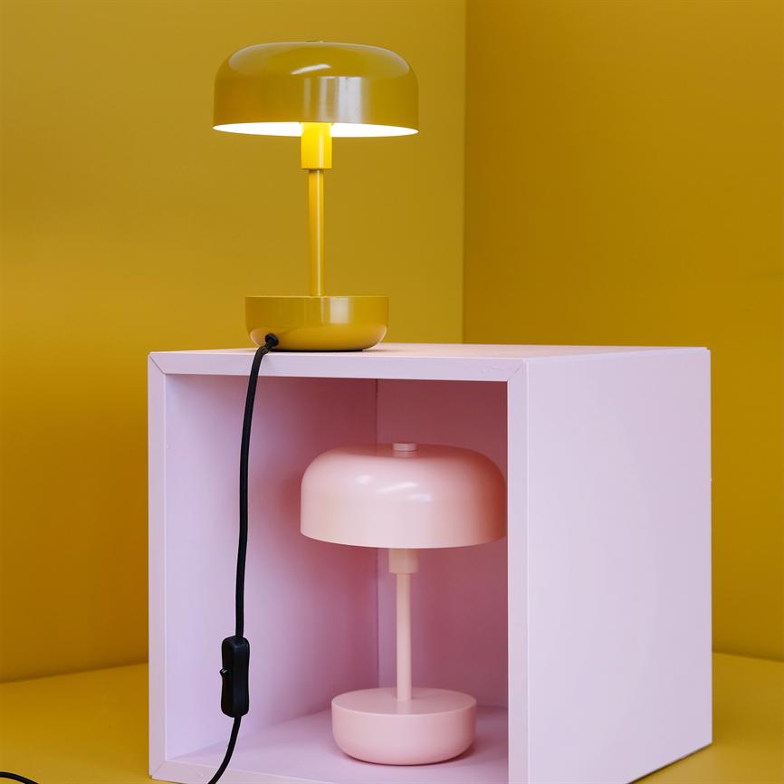Gul bordlampe lyser over en lyserød åben kasse, hvor en lyserød bordlampe står indeni; sort ledning med kontakt hænger ned foran en gul væg og bord.