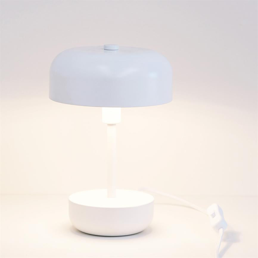 Hvid bordlampe lyser svagt, med rund skærm og cylindrisk fod; den står på en hvid flade foran en neutral, lys væg, med synlig strømledning og afbryder.