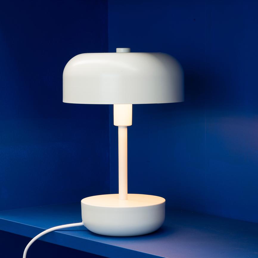 Hvid, rund bordlampe udsender blødt, varmt lys; den står på en blå hylde foran en dybblå væg.