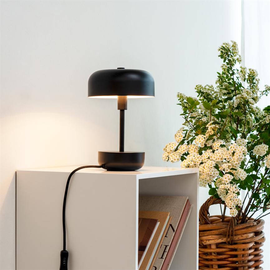 Sort bordlampe står tændt på et hvidt nattbord; ved siden af en flettet kurv med hvide blomster og en åben hylde med bøger, mod en lys væg.