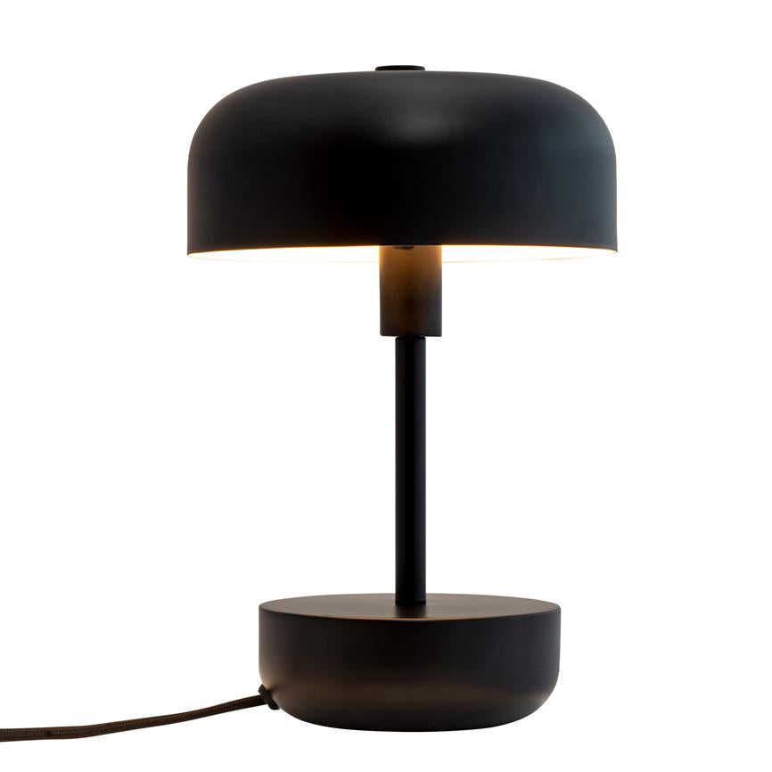 Sort bordlampe med rund skærm lyser nedad; slank cylinderformet stang forbinder skærm og bred, rund fod; stofbeklædt ledning strækker sig mod venstre på hvid baggrund.