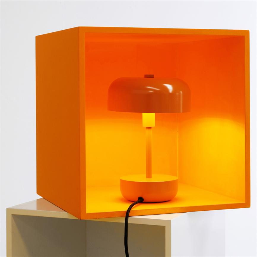 En orange bordlampe lyser inde i en åben orange kasse, placeret på en hvid hylde; en sort ledning hænger ud foran kassen.