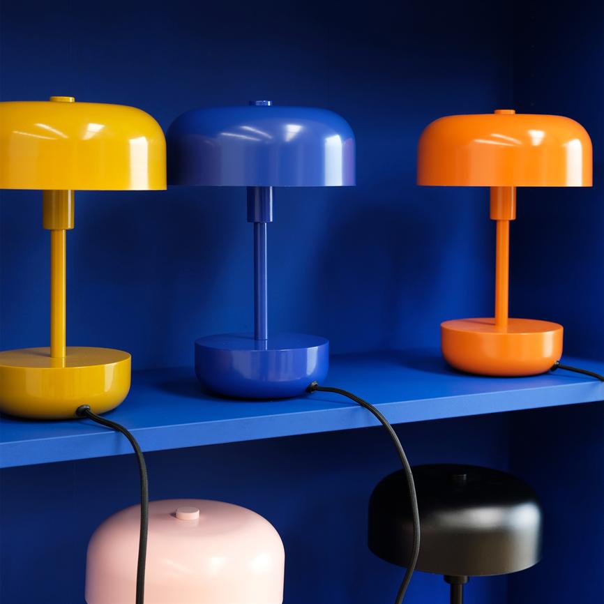 Tre bordlamper (gul, blå, orange) står på en blå hylde foran en blå væg; under ses en lyserød og en sort lampe på nederste hylde, med sorte ledninger hængende ned.