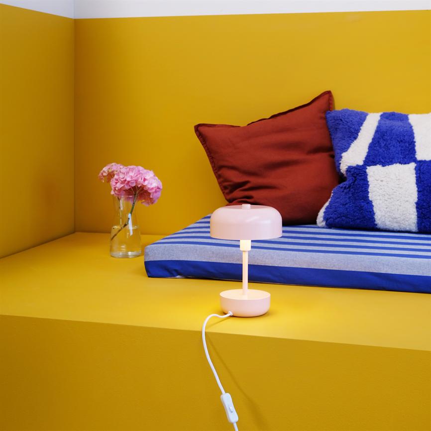 En lyserød bordlampe står tændt og kaster varmt lys på en gul platform; bagved ligger en blå-hvid stribet madras med rødt pudebetræk og en glasvase med lyserøde blomster.
