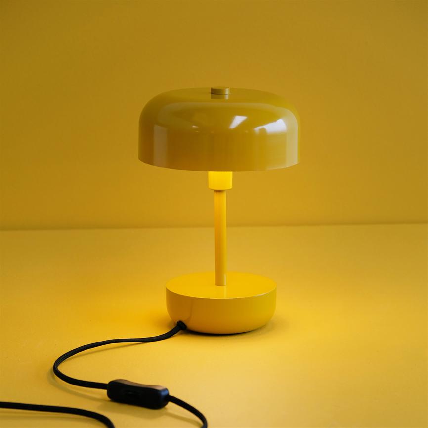 Gul bordlampe lyser blødt og kaster varmt lys nedad; står på gul overflade foran en gul væg, med en sort ledning og afbryder liggende foran.