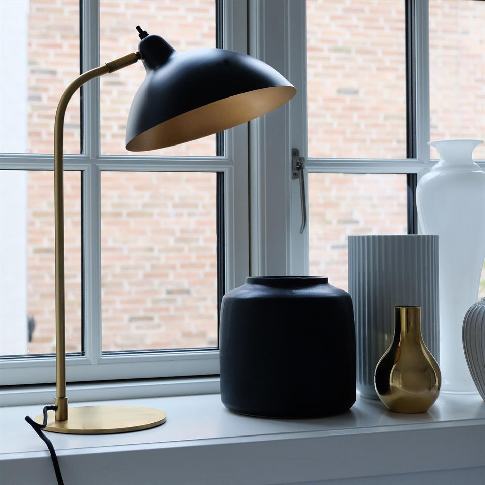 Sort og messingskrivebordslampe bøjer sig over vaser på en hvid vindueskarm; arrangement inkluderer en stor mat sort vase, ribbet hvid vase og lille guldfarvet vase; murstensfacade ses udenfor.