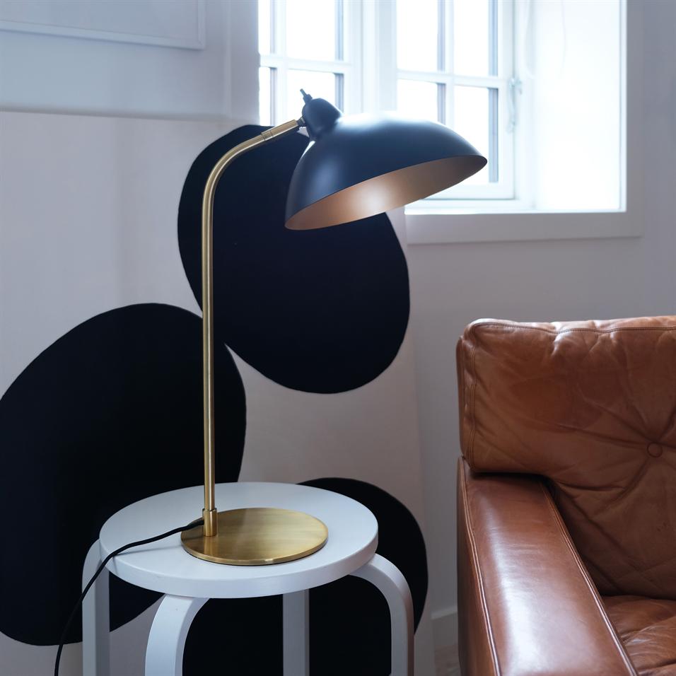 En sort bordlampe med gylden fod og vinklet skærm står på et rundt hvidt sidebord ved en lysebrun lædersofa foran et vindue og en væg med store sorte cirkler.