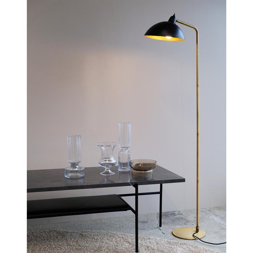 Gulvlampe med sort skærm og gyldent skaft kaster varmt lys over et mørkt marmorbord med fire glasvaser; står i et minimalistisk rum med lys væg, betongulv og lyst tæppe.