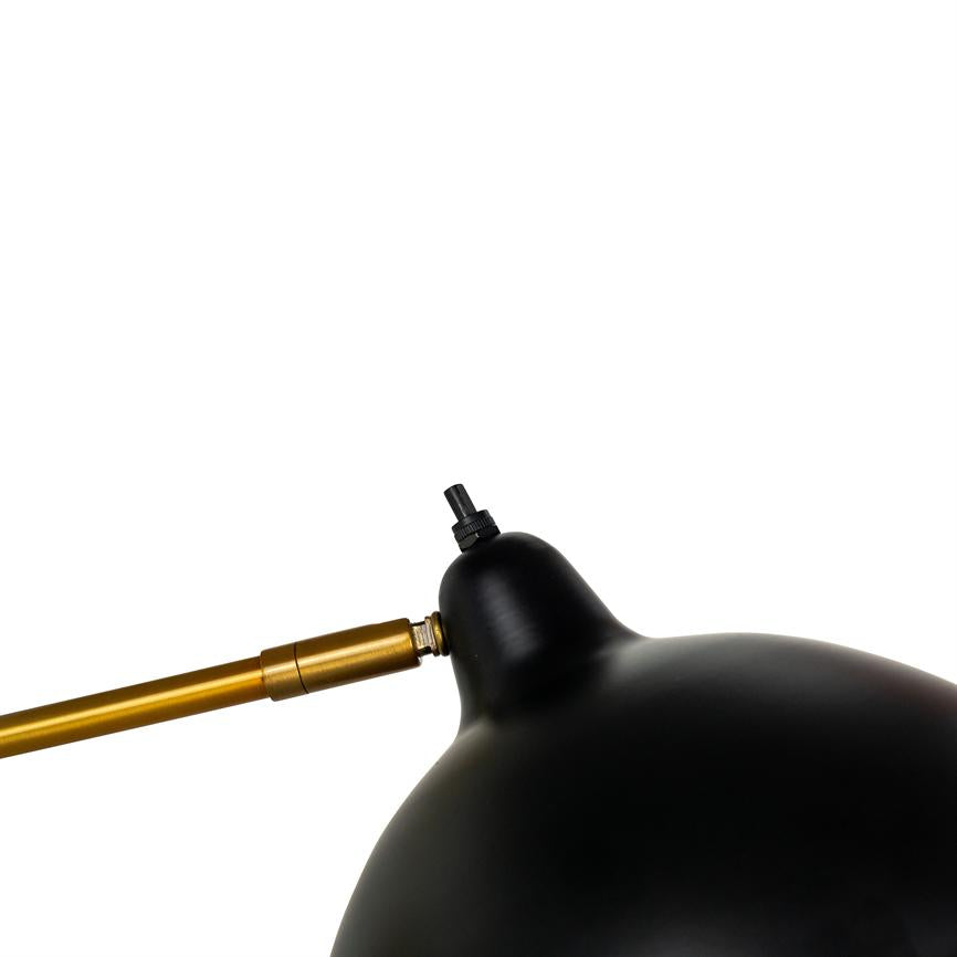 Sort metalskærm på en skrivebordslampe, forbundet til en guldfarvet arm med synligt hængsel og justerbar forbindelse, hælder i forgrunden mod en ensartet hvid baggrund.