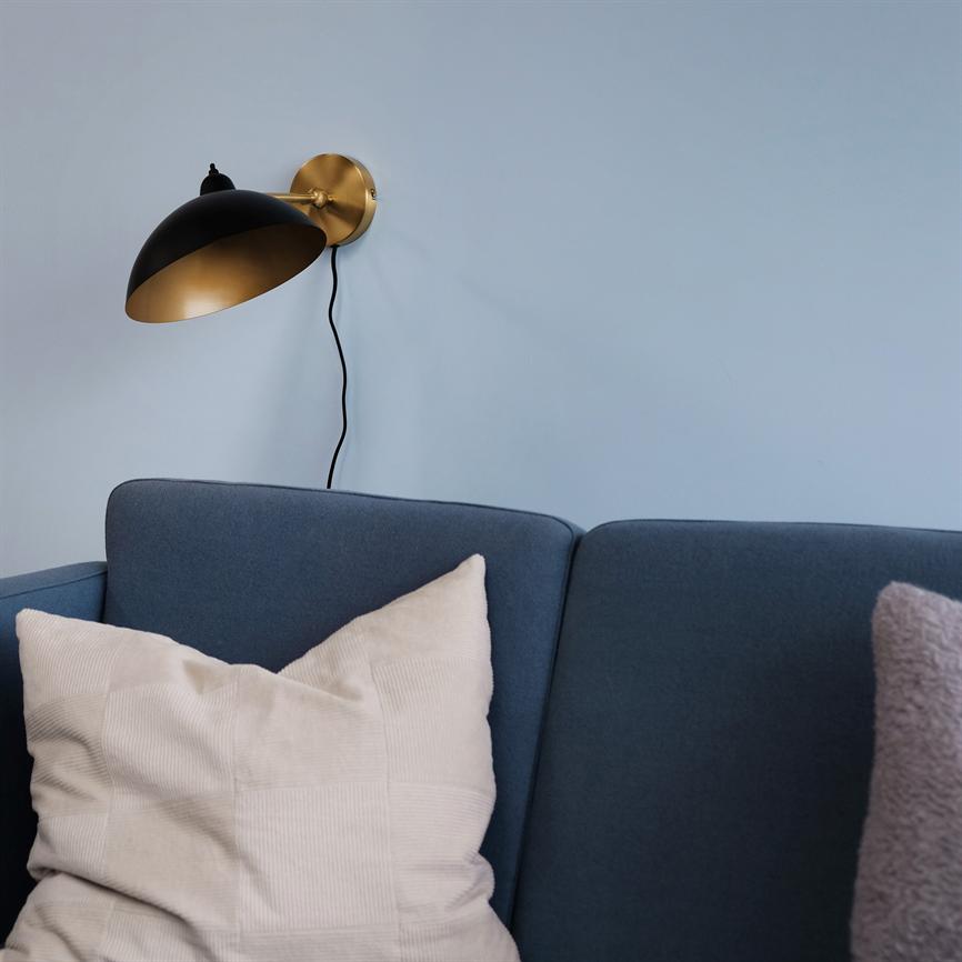 Sort- og guldfarvet væglampe monteret på en lyseblå væg, sort kabel hænger ned; lampen peger ned over en mørkeblå sofa med en stor lys pude og en lilla blød pude.