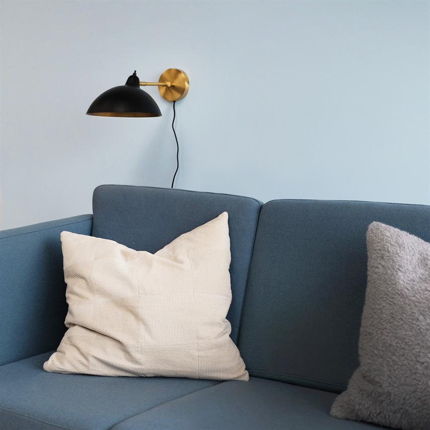 Blå sofa med en stor beige pyntepude ligger foran en lyseblå væg; en sort-guld væglampe hænger over sofaen, og en grå, fluffy pude ses i højre side.