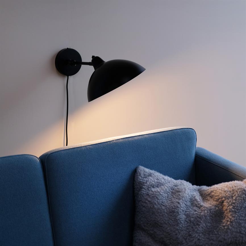 En sort væglampe kaster varmt lys over ryggen af en blå sofa, med en grå, blød pude på sædet i et roligt, neutralt stuemiljø.