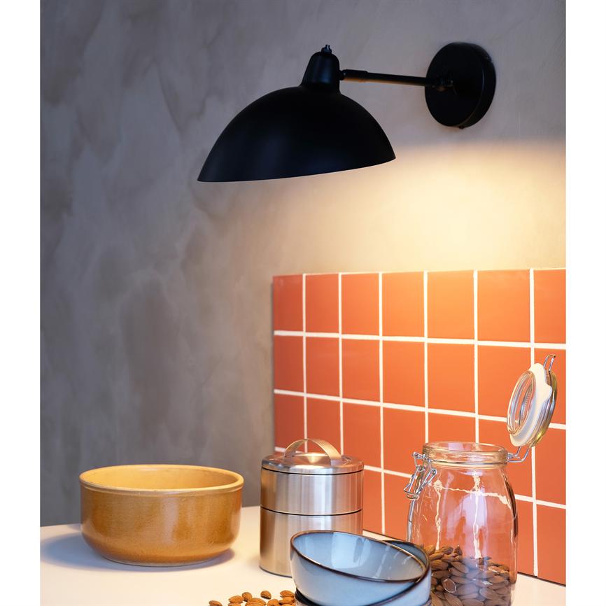 En sort væglampe lyser ned over et rødt flisebagpanel og et køkkenbord med keramisk skål, metalbeholder, åben glaskrukke og spredte mandler.