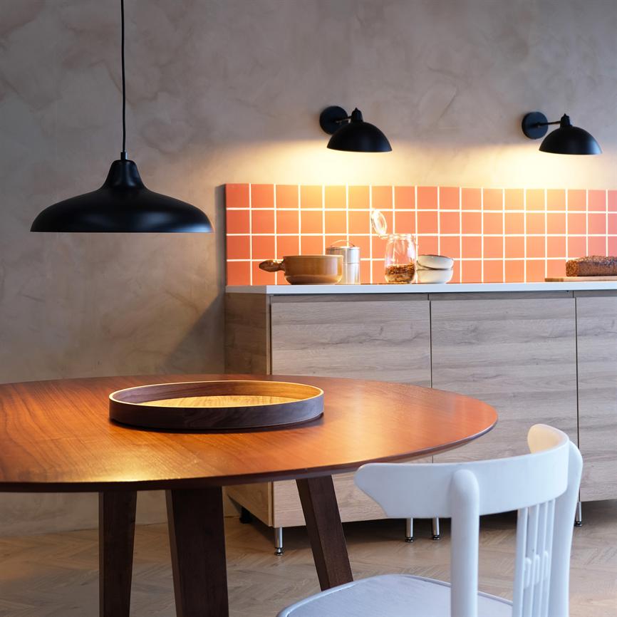 Rundt træbord med træbakke står foran en hvid stol, oplyst af en sort pendel og væglamper; bagved køkkenbord med lyse træskabe, orange fliset backsplash, krukker, pande og brød.