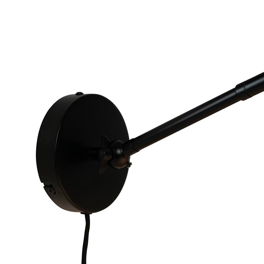 Sort rund vægbeslag holder en lang, slank lampearm, som strækker sig mod højre; sort stofledning hænger ned, alt mod en neutral hvid baggrund.