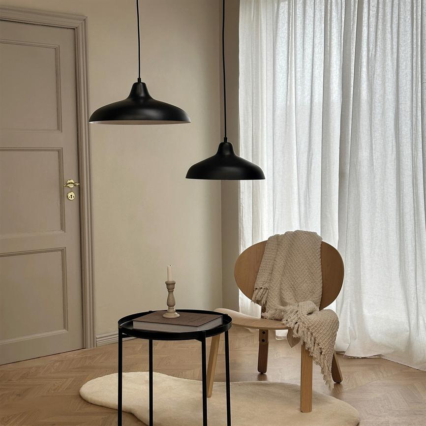En træstol bærer et beige, vævet tæppe, placeret ved et rundt sort sidebord med en enkelt lysestage; to sorte pendellamper hænger over stolen, foran lyse gardiner og trægulv.