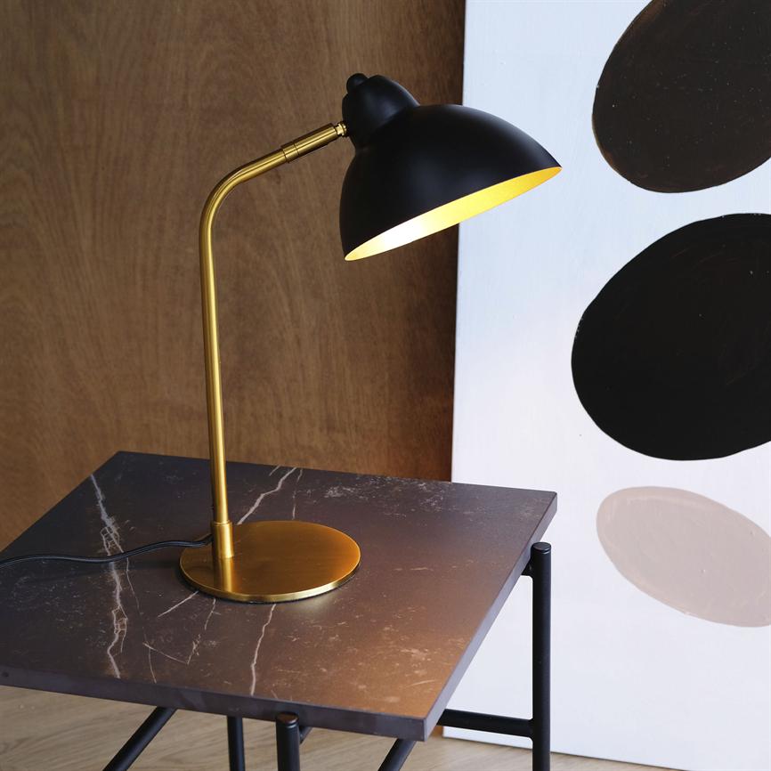 Objekt: Sort bordlampe med gylden stang og fod; Handling: lyser og kaster varmt lys nedad; Kontekst: på mørkt marmorbord foran trævæg og et lærred med sorte og beige cirkler.