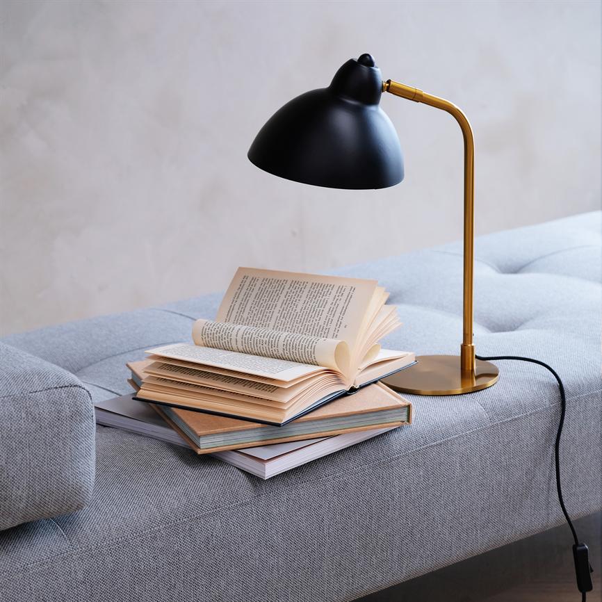 En sort og guldfarvet læselampe oplyser en åben bog, der ligger på en stak bøger på en grå sofa i et lyst, minimalistisk rum; ingen læsbar tekst at transskribere.