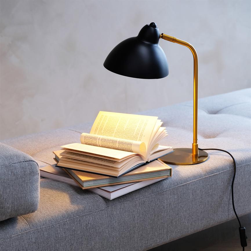 Sort og guldfarvet bordlampe lyser over en stak åbne bøger, der ligger på en lysegrå sofa i et roligt, neutralt rum.