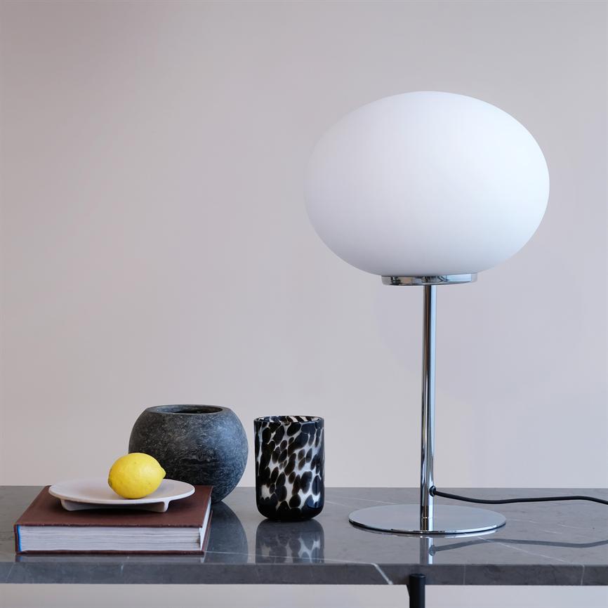 En hvid rund bordlampe står på et kromstativ på en mørk marmorbordplade; ved siden af en bog med en tallerken og citron og to vaser foran en beige væg.