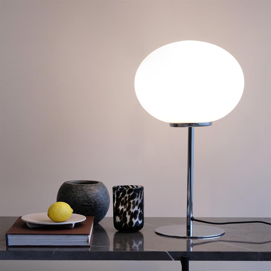 Bordlampe med rund, hvid glas-skærm lyser blødt på et marmorbord, flankeret af en bog med en tallerken og citron, en mørk stenurne og et sort-hvidt mønstret glas.