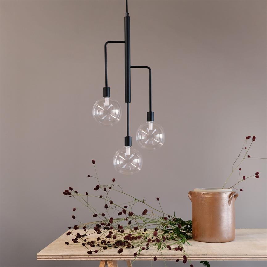 En moderne pendel med tre klare glaskupler hænger over et bord, hvor vilde rødbrune blomster ligger spredt, og en brun keramikkrukke står mod en støvet rosa væg.