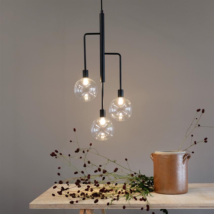 En sort pendel med tre oplyste glaskugler hænger over et lyst bord med en brun keramikkrukke og spredte tørrede, mørkerøde blomster mod en beige væg.